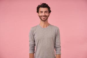 Studio shot of beautiful bearded male with dark hair standing over pink background, wearing casual clothes, looking at camera cheerfully with hands down photo