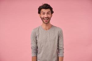 Indoor portrait of happy bearded male with trendy haircut wearing casual clothes, posing over pink background with hands down, looking at camera cheerfully with wide mouth opened photo