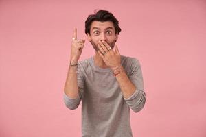 Surprised pretty bearded male with trendy haircut wearing grey sweater, standing over pink background with palm on his mouth, looking at camera and pointing upwards with index finger photo