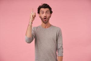 Portrait of dark haired bearded male posing over pink background with amazed face, wearing grey sweater, looking at camera and contracting forehead, pointing upwards with index finger photo