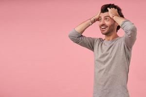 Handsome shocked bearded young man with dark hair wearing grey sweater, standing over pink background with wide eyes opened, looking aside and holding head with hands like he do not believe in news photo