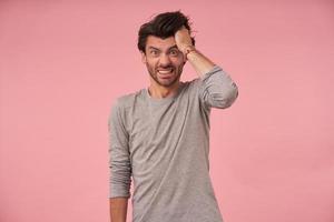 Studio portrait of stressed bearded man with dark hair posing over pink background, wearing casual clothes, looking at camera and showing teeth, holding head with hand and rumpling photo