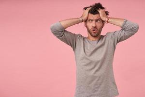 Depressed young beautiful man with beard standing over pink background with palms on his head, looking aside with desperate face, frowning and contracting forehead photo