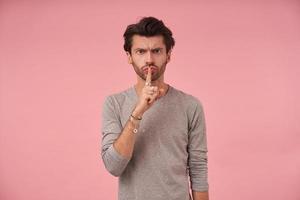 Young dark haired man with beard looking at camera with forefinger on his mouth, asking to keep silence with serious face, standing over pink background in casual clothes photo