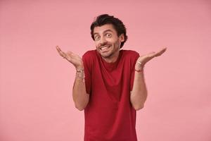 Indoor shot of funny young bearded male with dark hair standing over pink background with raised palms, looking at camera cheerfully and shrugging, contracting forehead and smiling broadly photo