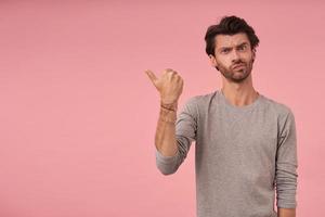 retrato de estudio de un joven barbudo serio con suéter gris parado sobre fondo rosa, mirando la cámara y frunciendo el ceño, señalando a un lado con el pulgar levantado foto