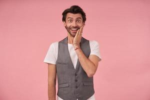 Indoor photo of joyful bearded male with trendy haircut looking at camera with wide smile, wearing grey waistcoat and white t-shirt, standing over pink background and keeping palm on his cheek