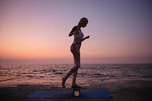 Outdoor photo of sporty slim female with casual hairstyle in sporty wear standing on balance board, doing sports with balancer on seafront during sunrise, raising hands to save equilibration