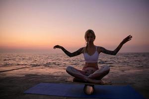 Young healthy blonde woman in good physical condition sitting on balance board with crossed legs, wearing sporty clothes, balancing on board over seaside view during sunrise with raised hands photo