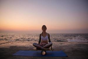 Outdoor photo of slim blonde woman sitting on balance board with closed eyes and calm face, posing over sea view during sunrise, helping to keep balance with hands
