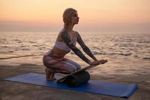Outdoor shot of blond sporty woman with body in good physical condition posing over sea view, making sports on early morning with mat and balance board, wearing sporty top and leggins photo