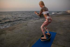 Young sporty woman with blonde hair doing exercise with balancer on seafront, standing on special sports equipment and trying to keep balance, wearing sport clothes photo