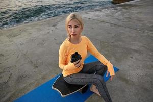 Outdoor shot of young pretty blonde lady with casual hairstyle wearing orange long sleeve top and leggins doing, posing over seaside on early morning, holding shaker in hand and looking aside photo