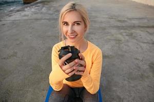 Outdoor close-up of positive young woman with blonde hair posing over seaside with shaker in her hands, looking at camera with sincere light smile photo