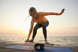 Outdoor shot of young lovely woman trying to keep balance on wooden desk, balancing over sea view during sunrise, wearing orange long sleeve top and dark leggins photo