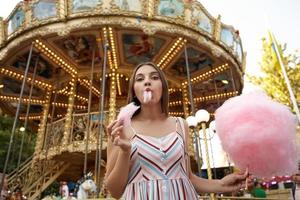 Una joven guapa de buen aspecto con el pelo castaño y un vestido romántico parada sobre un carrusel en un parque de atracciones, comiendo algodón de azúcar en un palo de madera foto