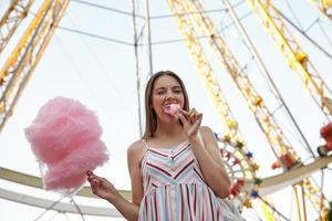 mujer hermosa joven positiva en vestido de verano de pie sobre la rueda de la fortuna en el parque de atracciones en un día cálido, sosteniendo algodón de azúcar en un palo y poniéndose un trozo en la boca, guiñando un ojo a la cámara foto