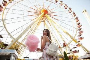 Outdoor shot of happy young brunette lady with long hair wearing romantic dress and white backpack, standing over ferris wheel on summer warm day, holding cotton candy and smiling widely photo