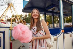 Posititve young attractive brunette female with long hair posing over amusement park on warm sunny day, wearing romantic dress and backpack, keeping in hand stick with pink cotton candy