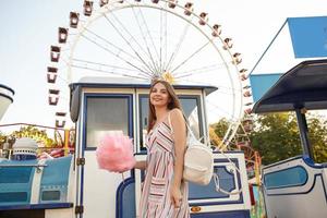 Encantadora joven morena con el pelo largo y castaño posando sobre la rueda de la fortuna en un día cálido y soleado, con un vestido romántico y una mochila blanca, sosteniendo algodón de azúcar en un palo foto