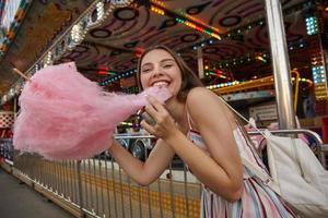 Portrait of happy young pretty woman with brown hair wearing light summer dress, walking through amusement park on warm day, holding cotton candy in hand and pulling it with teeth photo