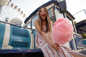 Outdoor photo of pretty joyful female with long hair sitting on steam train over amusement park, holding cotton candy in hand and looking cheerfully to camera with wide mouth opened