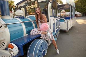 alegre mujer morena de pelo largo con vestido ligero de verano sentada en un vagón de tren de vapor en un parque de atracciones en un día soleado y cálido, sosteniendo algodón de azúcar en un palo y mirando y cámara felizmente foto