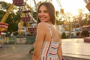 Outdoor back view of young pretty female with brown hair posing over amusement park on warm summer day, turning around and looking to camera happily with charming smile
