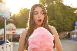 Outdoor shot of beautiful young brunette long haired woman posing over green park on warm sunny day, looking on pink cotton candy with surprised face and opening mouth widely photo