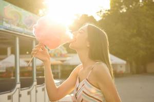 Outdoor sunny shot of happy pretty long haired lady in romantic dress posing over amusement park on warm summer day, eating pink cotton candy with big pleasure photo