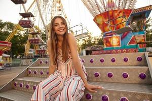 Outdoor photo of charming young lady in romantic summer dress sitting over amusement park decorations, being in nice mood, looking to camera with wide sincere smile