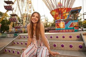 Charming cheerful young long haired brunette female posing over amusement park on warm sunny day, wearing romantic dress with straps, smiling to camera happily photo