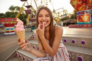 joven y alegre mujer hermosa con el pelo castaño largo sentada en el parque de atracciones con helado en la mano, mirando a la cámara con una amplia sonrisa y manteniendo el pulgar en el labio inferior foto