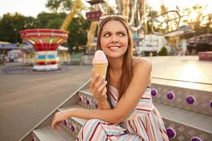 Puzzled young long haired female with sunglasses on her head looking aside and frowning, holding ice cream cone in hand while sitting over amusement park decorations photo