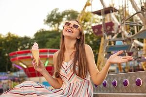 Happy young beautiful woman in romantic light dress sitting over amusement park decorations with ice cream cone in hand, smiling with closed eyes and raising palm up