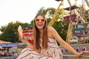 Outdoor photo of joyful young long haired brunette female in sunglasses sitting over amusemant park and leaning on stairs with hand, laughing happily and holding ice cream in hand