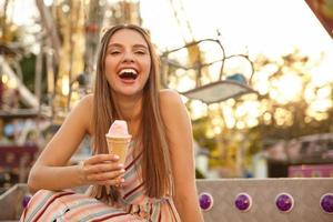 Portrait of joyful young lovely woman with sunglasses on her head posing outdoor over park of attractions, laughing with wide mouth opened and keeping ice cream cone in hand photo