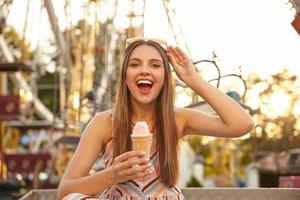 Young cheerful long haired pretty lady with sunglasses on her head looking at camera with broad smile, eating ice cream cone while walking through park of attractions, wearing summer clothes photo