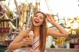 Outdoor shot of pretty young happy female with long hair eating ice cream in cone while walking through amusement park, laughing joyfully with wide mouth opened and taking sunglasses off photo