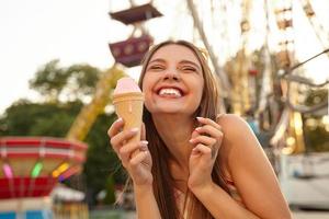 Close-up of charming cheerful young brunette lady with sunglasses on her head, showing teeth and smiling happily while eating pink ice cream cone over amusement park