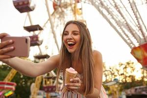 Portrait of happy charming young brunette female wearing summer light dress, making selfie outdoor with her smartphone, keeping ice cream cone in hand and smiling joyfully photo