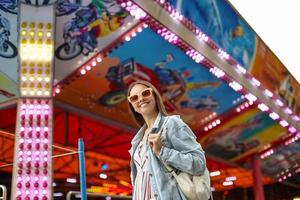 foto al aire libre de una hermosa joven con gafas de sol caminando por un parque de diversiones en un día cálido, usando un vestido romántico y un abrigo de jeans, sonriendo sinceramente a la cámara