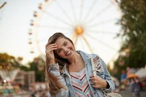 Outdoor photo of beautiful brunette female straightening hair and looking to camera with charming wide smile, standing over ferris wheel on warm bright day in casual clothes