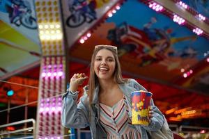 atractiva joven morena con abrigo de jeans y gafas de sol en la cabeza sosteniendo palomitas de maíz en la mano y tirándolas a la cámara, de pie sobre las atracciones en el parque de diversiones foto