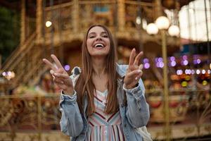 foto al aire libre de una hermosa joven con el pelo largo y castaño con un vestido romántico y un abrigo de jeans, de pie sobre el carrusel en el parque de atracciones, sonriendo a la cámara y levantando los dedos en señal de victoria