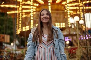 mujer joven y bonita con el pelo largo posando sobre atracciones en el parque de atracciones, vestida con vestido de verano y abrigo de jeans, mirando a la cámara y sonriendo sinceramente foto