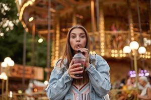 Outdoor photo of lovely long haired brunette woman in casual clothes drinking lemonade with straw while walking in park of attractions, looking at camera and contracting forehead