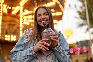 Positive long haired young pretty female in trendy jeans coat posing over park of attractions, giving wink and smiling widely to camera while drinking lemonade with straw photo