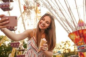 Beautiful young long haired brunette lady in romantic summer dress posing over attractions while making selfie with her mobile phone, keeping ice cream cone in hand and smiling cheerfully to camera photo