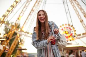 Outdoor shot of joyful long haired young pretty female in romantic dress and jeans coat posing over ferris wheel, keeping cup of lemonade in hands and smiling cheerfully to camera photo